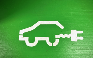 Zöld autó festett ikonon való ábrázolása: fehér autó töltőkábellel, zöldre festett falon.